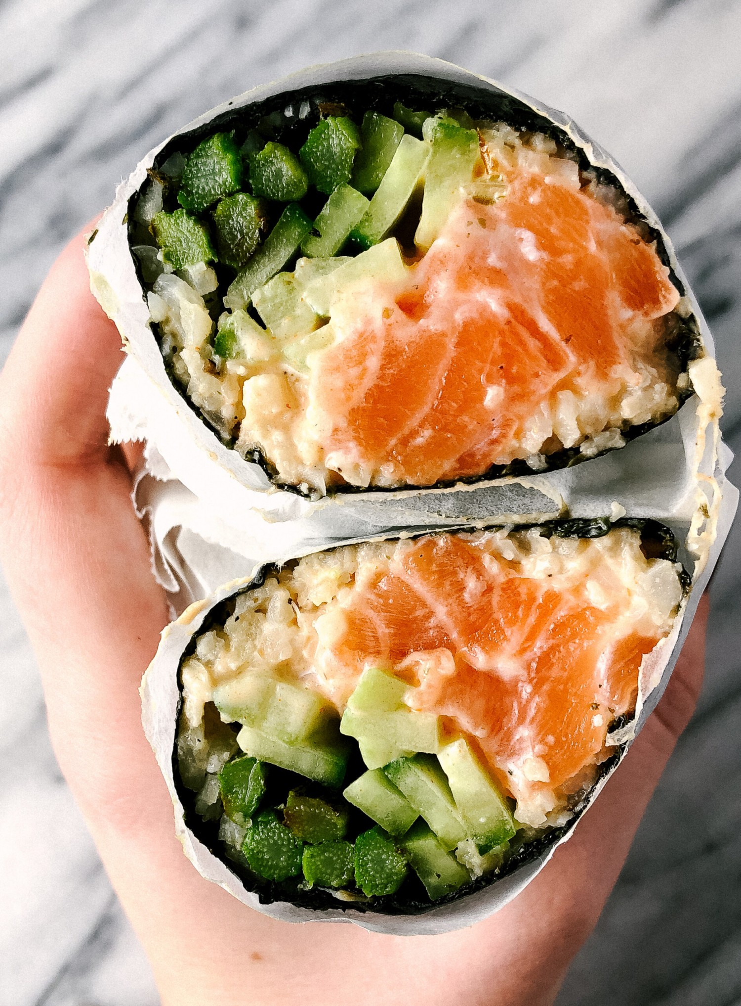 Side view of a sushi burrito cut in half. There is rice, raw salmon, veggies, and spicy mayo all wrapped in a sheet of nore