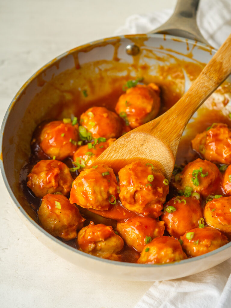 Three quarter view of sweet and sour meatballs in a large frying pan with a wooden spoon