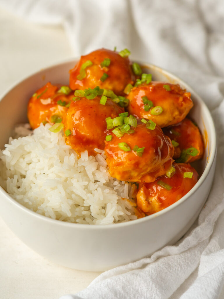 Three quarter view of sweet and sour chicken meatballs in a serving dish with rice and garnished with green onion