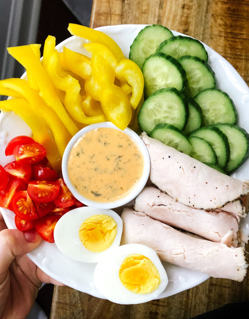 A snack plate with cut bell pepper, cucumber, tomatoes, turkey lunch meat, hard boiled egg and a dipping sauce