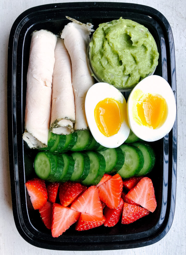 Black tupperware container with lunchmeat, mashed avocado, jammy egg, sliced cucumber an strawberries inside