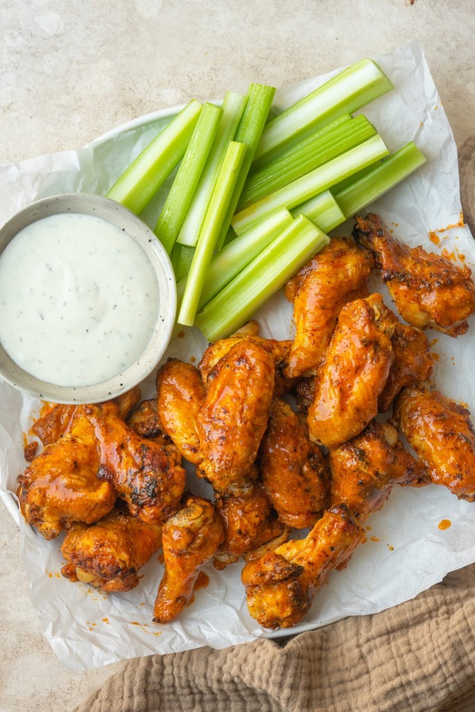 Air fryer buffalo wings on a plate lined with parchment paper. The wings are next to celery and ranch dressing in a small dish