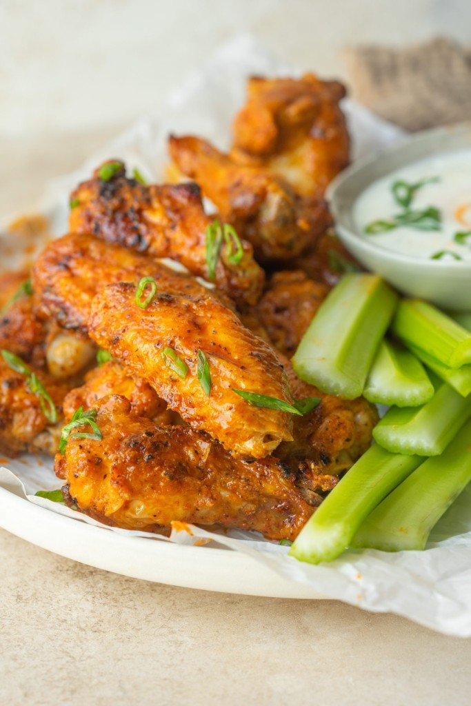 Three quarter view of buffalo wings stacked on a serving plate next to celery and ranch for dipping