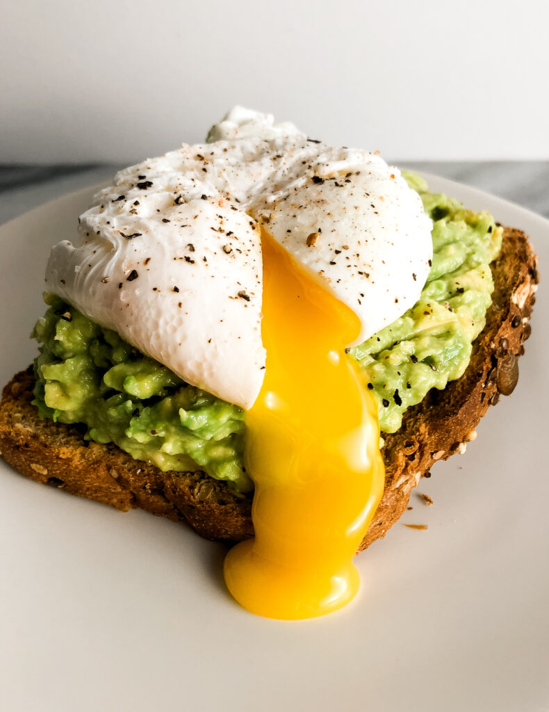 three quarter view of  a perfect poached egg on a piece of avocado toast. The poached egg is sliced open and yolk is coming out.