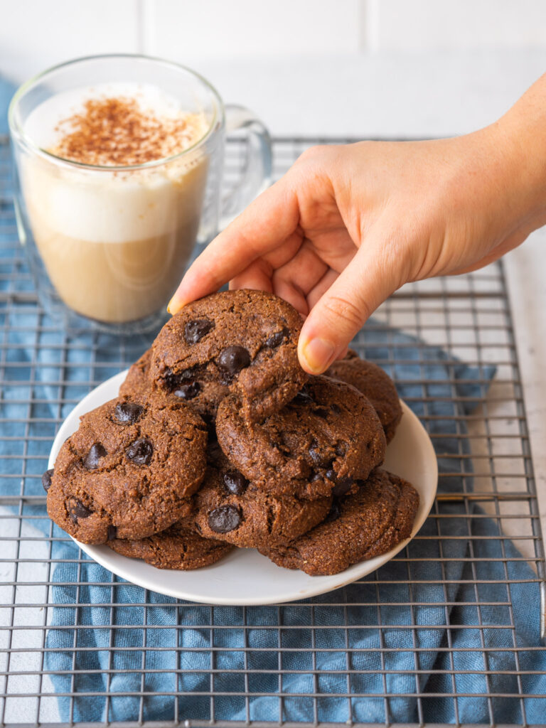 hand grabbing chocolate chip mocha cookies off a plate with a mocha latte in the back