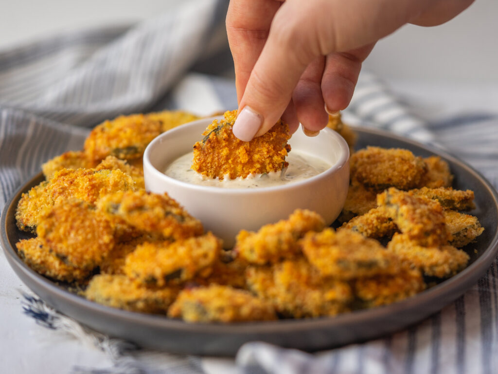 side view of a hand holding a low carb fried pickle dipping into ranch surrounded by air fryer pickles