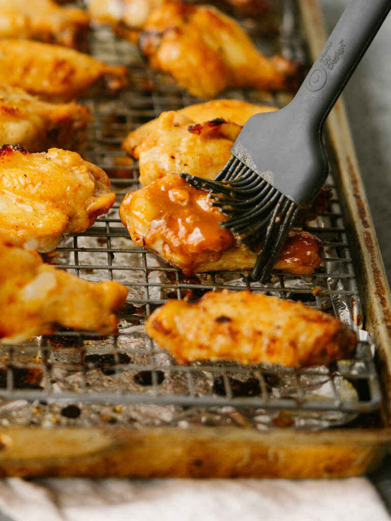 a close up photo of a grill brush brushing sauce on a chicken wing on a baking sheet