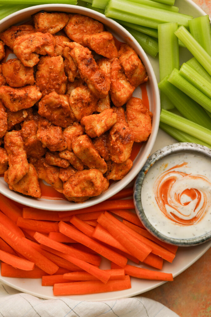 an above view photo of buffalo chicken bites in a bowl surrounded by celery and carrots and a small bowl of ranch dressing