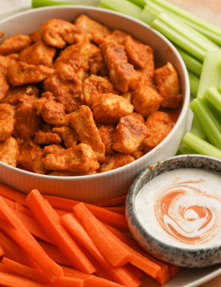 an three quarter view photo of buffalo chicken bites in a bowl surrounded by celery and carrots and a small bowl of ranch dressing on a plate