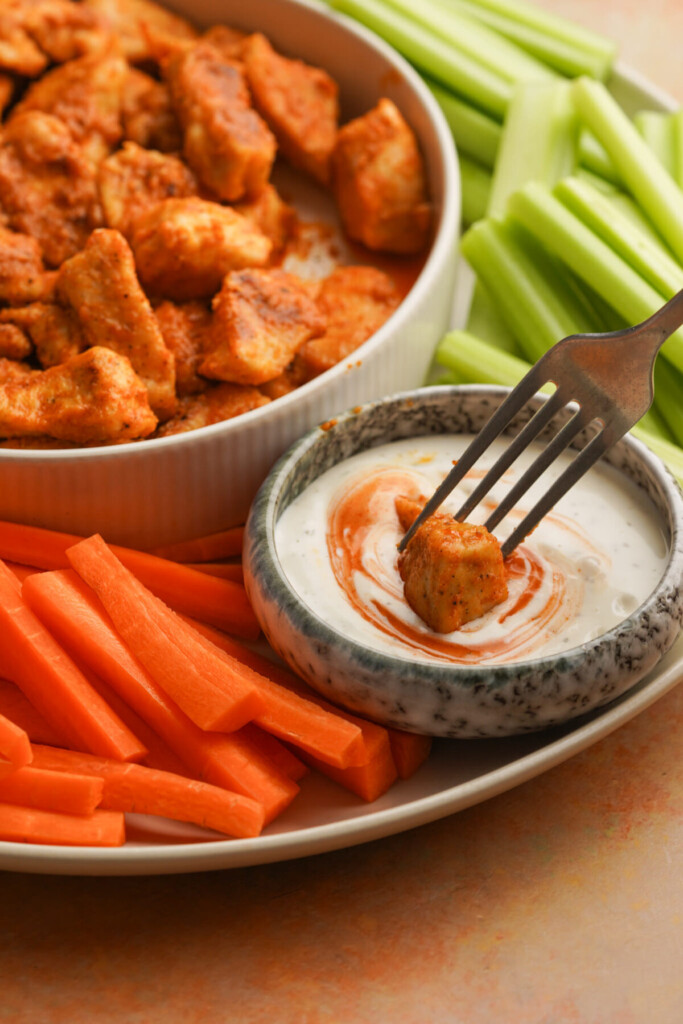 a side view photo of a fork with a buffalo chicken bite on it dipping into a small bowl of ranch with carrots and celery next to it