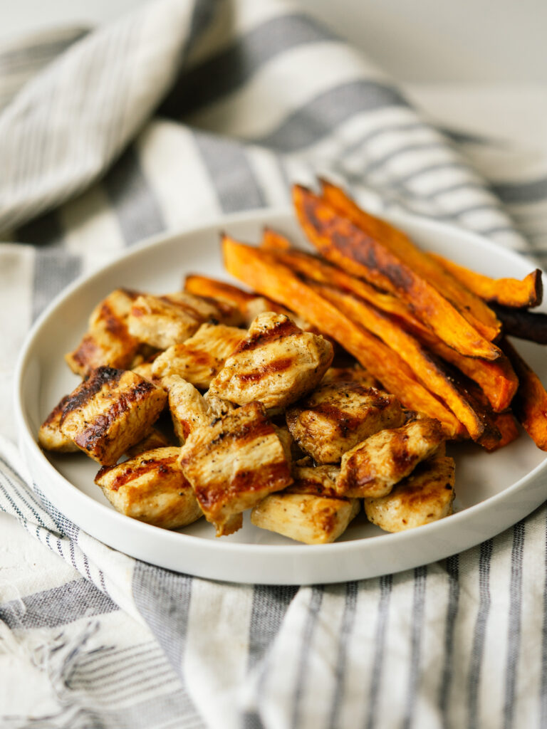 a three quarter view photo of grilled chicken nuggets on a white plate with sweet potato fries