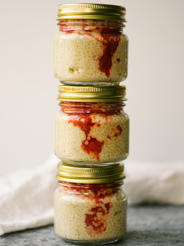 Side view of glass jars with gold metal lids stacked on top of each other. There is mango chia seed pudding with strawberry compote in each jar.