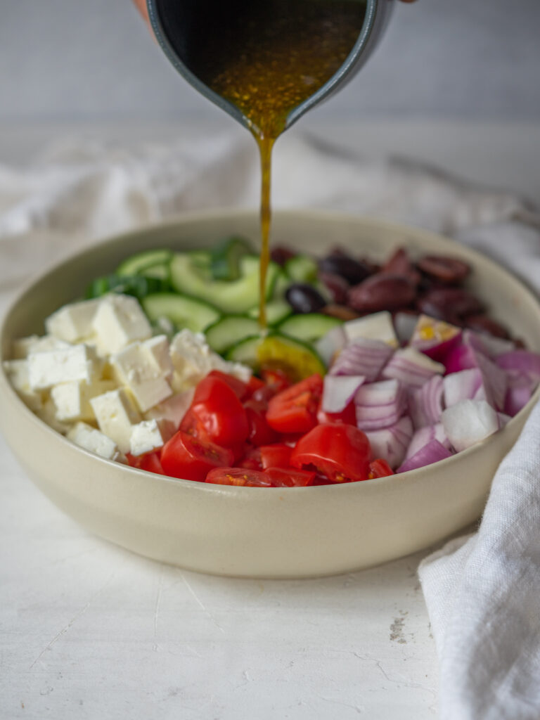 Greek salad dressing pouring onto chopped greek salad ingredients in a bowl