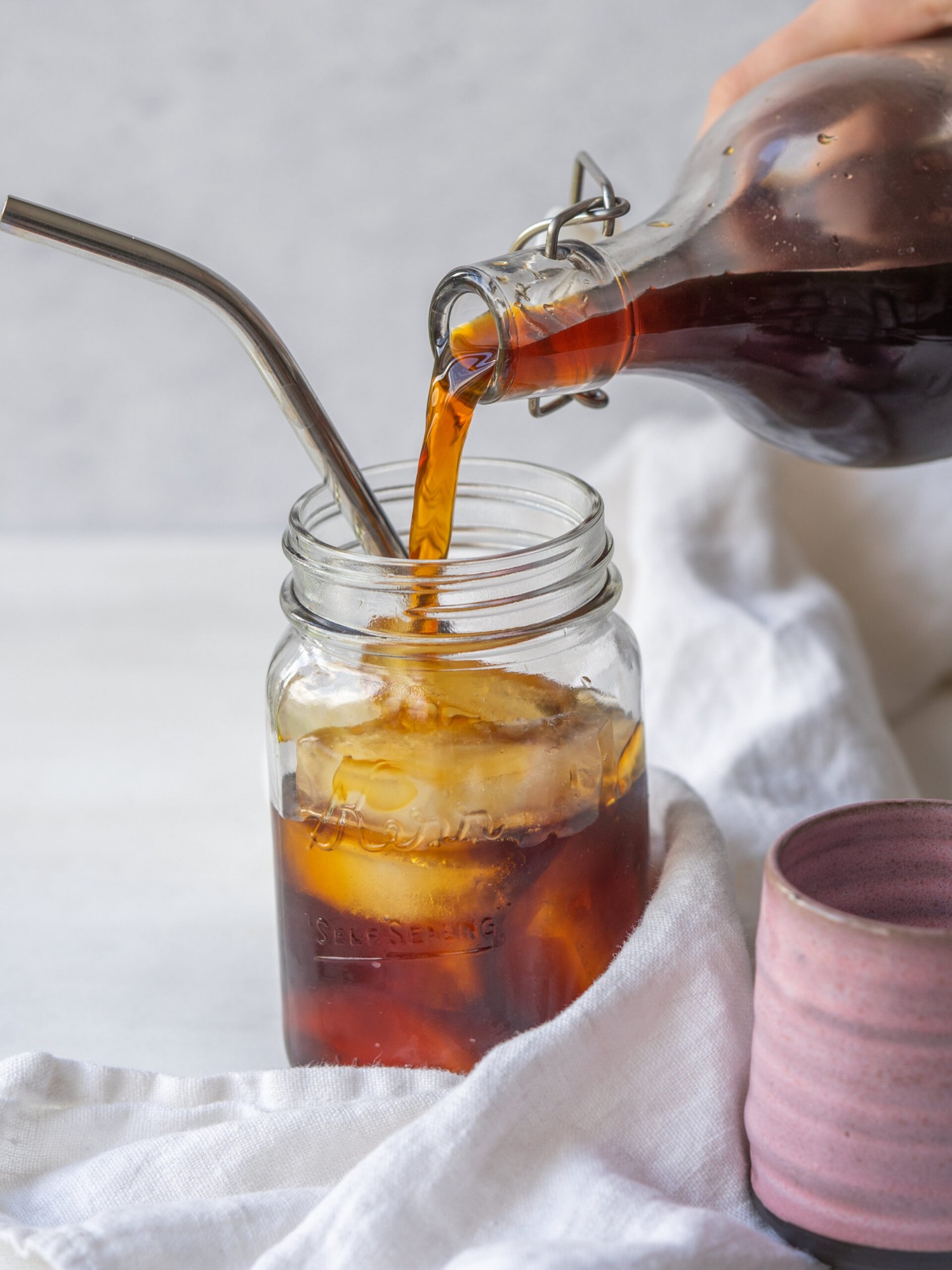 a side view photo of a glass container pouring cold brew into a mason jar with ice and a straw in it