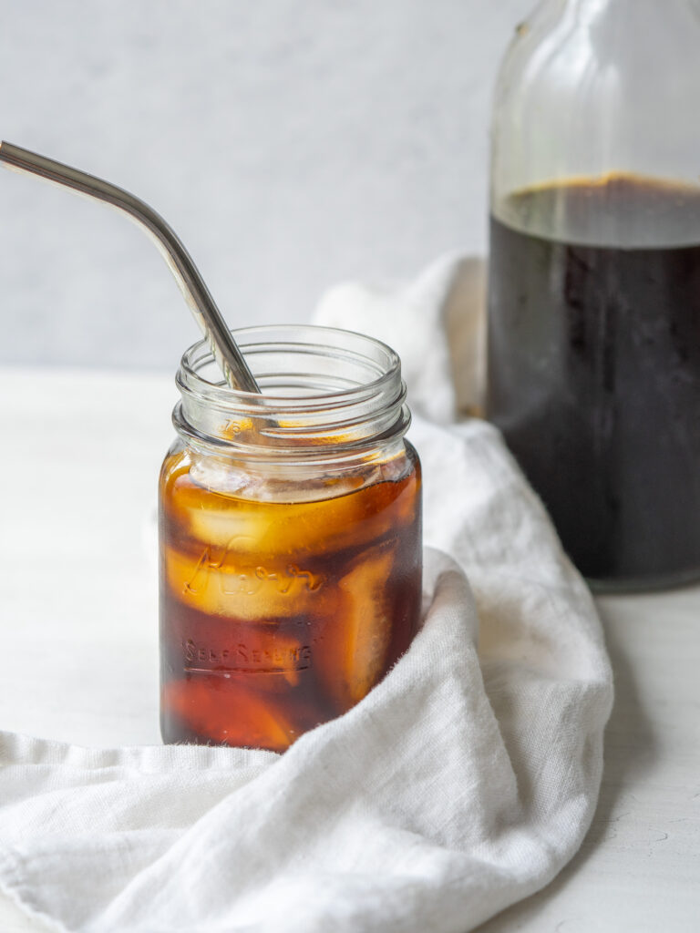 a side view photo of cold brew in a mason jar with ice and a straw and a glass jug of cold brew behind it