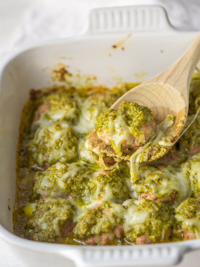 Three quarter view of a wooden spoon scooping turkey pesto meatballs out of the baking dish