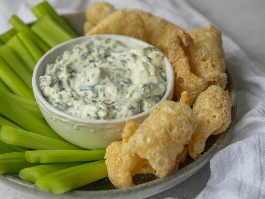 Three quarter view of healthy spinach dip on a serving plate with celery and pork rinds