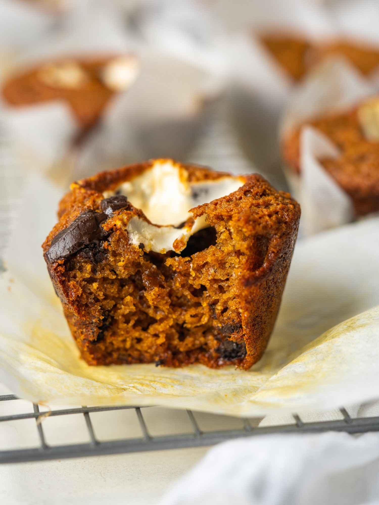 a side view photo of a s'mores muffin with a bite taken out of it on parchment paper on a wire rack
