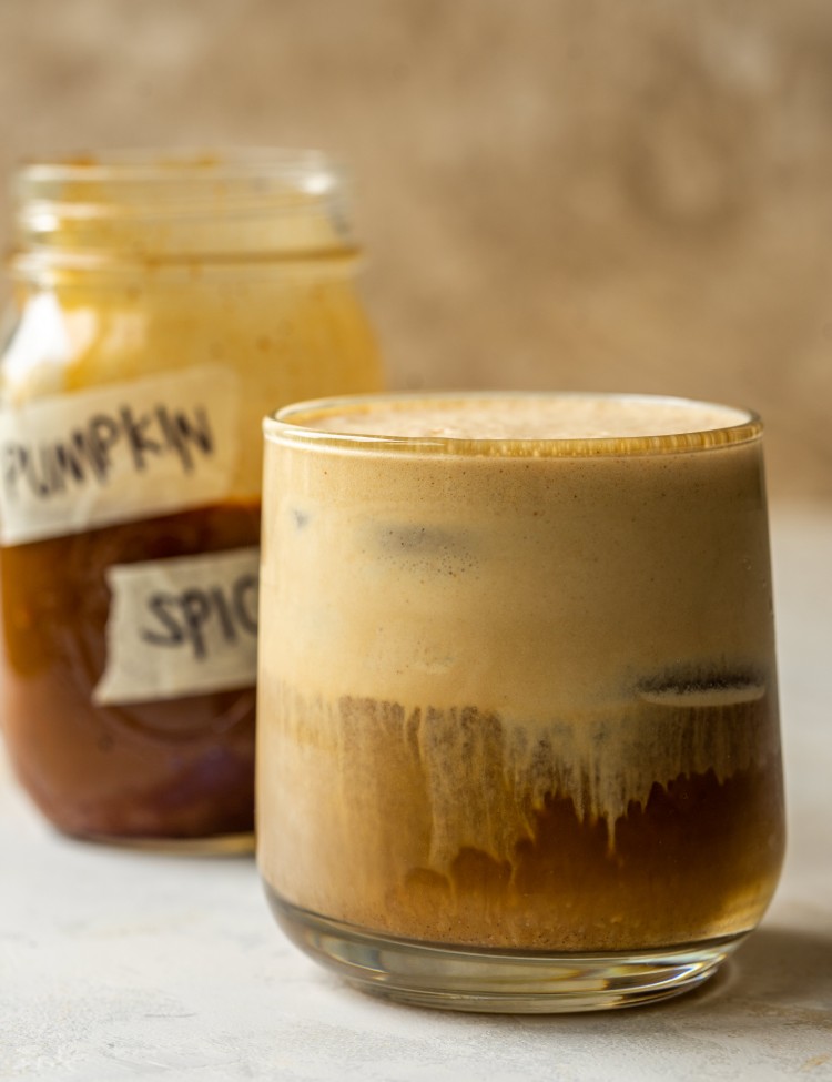 Side view of a pumpkin cream cold brew drink next to a jar of pumpkin spice syrup