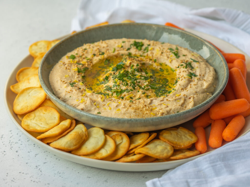 Side view of a creamy hummus recipe in a bowl served with carrots and pita crisps