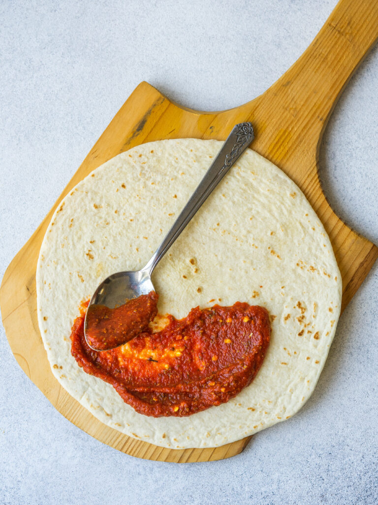 Above view of a spoon spreading sauce over a tortilla to make a tortilla pizza