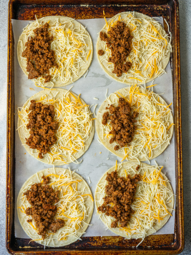 Above view of white corn tortillas on a sheet pan topped with shredded cheese and ground beef