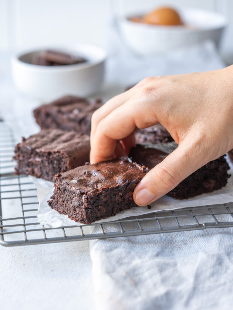 Side view of a hand grabbing a flourless double chocolate brownie off of  a cooling rack