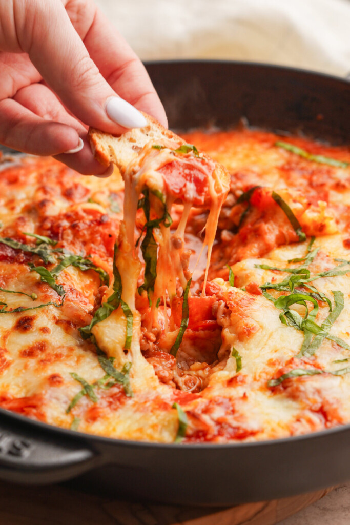 a close up photo of a hand holding a crostini with pizza dip on top above the pizza dip in a cast iron skillet