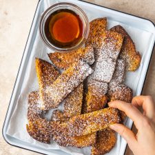 an above picture of a hand holding a french toast stick over a baking sheet filled with french toast sticks and dusted with powdered sugar