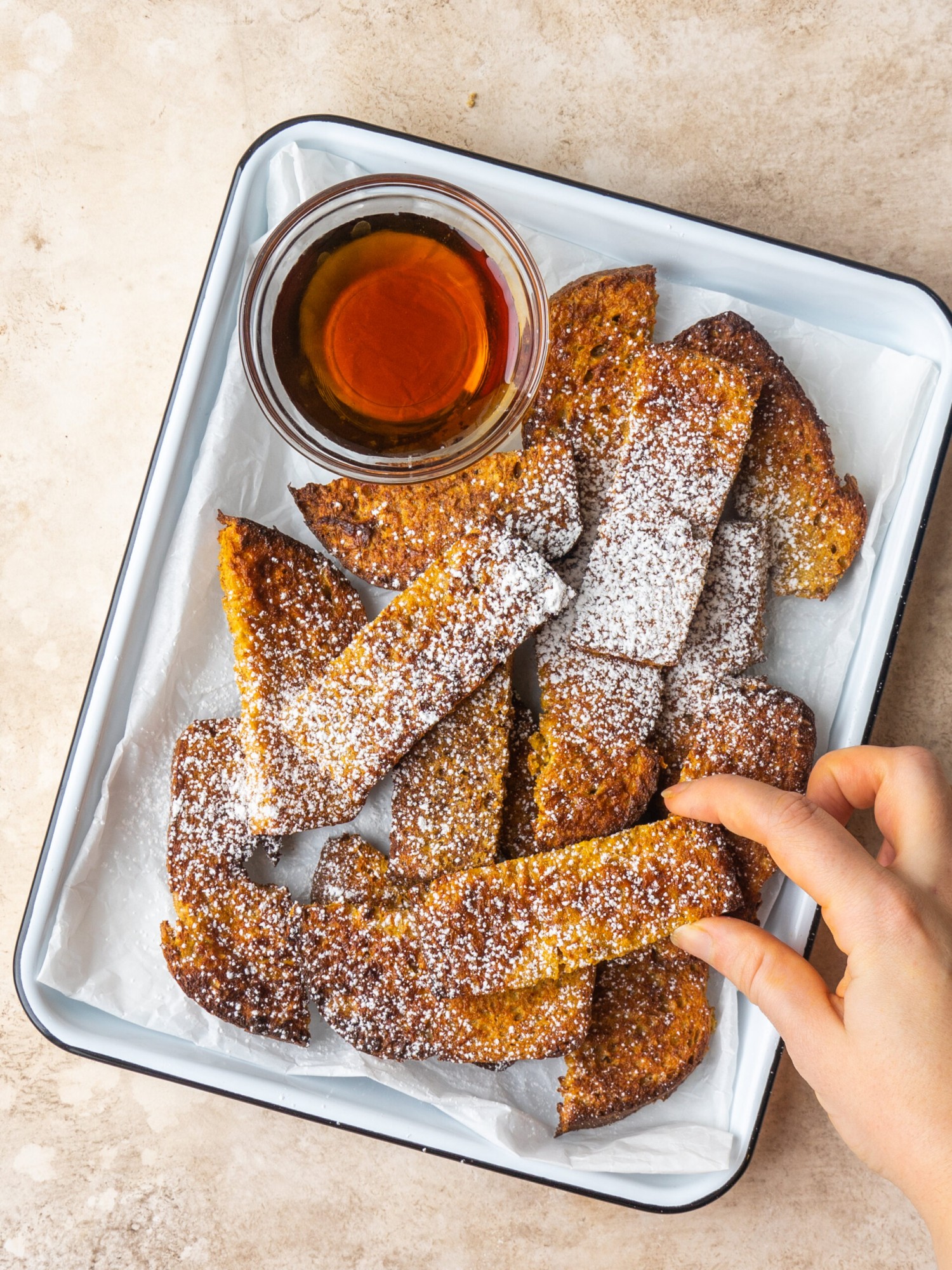 an above picture of a hand holding a french toast stick over a baking sheet filled with french toast sticks and dusted with powdered sugar
