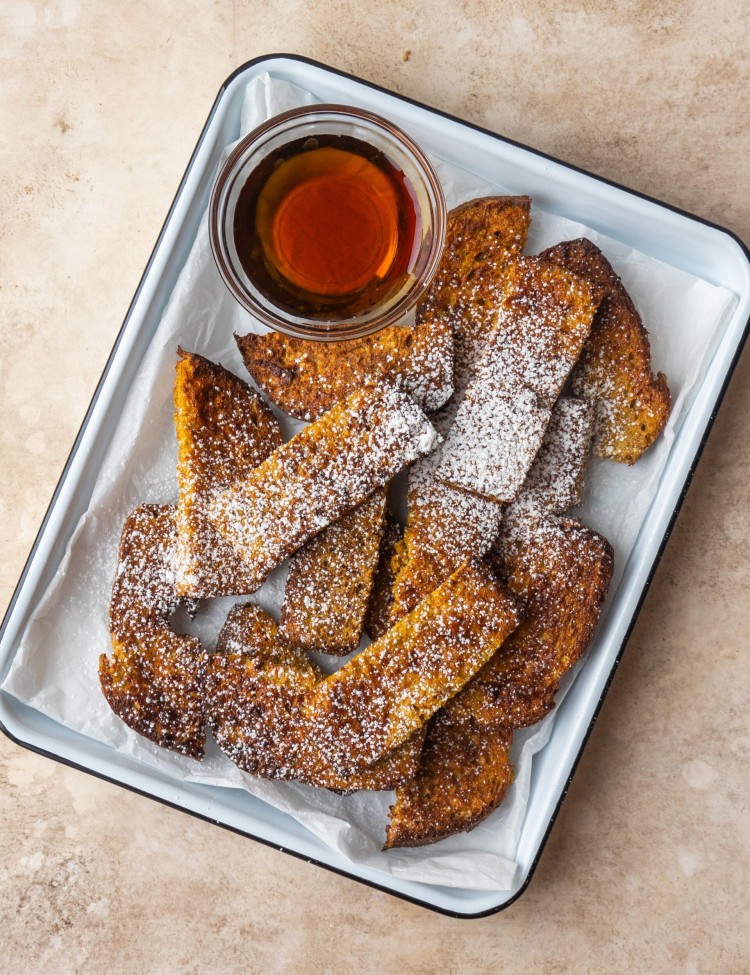 an above image of air fryer french toasts sticks on a baking sheet lined with parchment paper with powdered sugar on top and a small bowl of maple syrup
