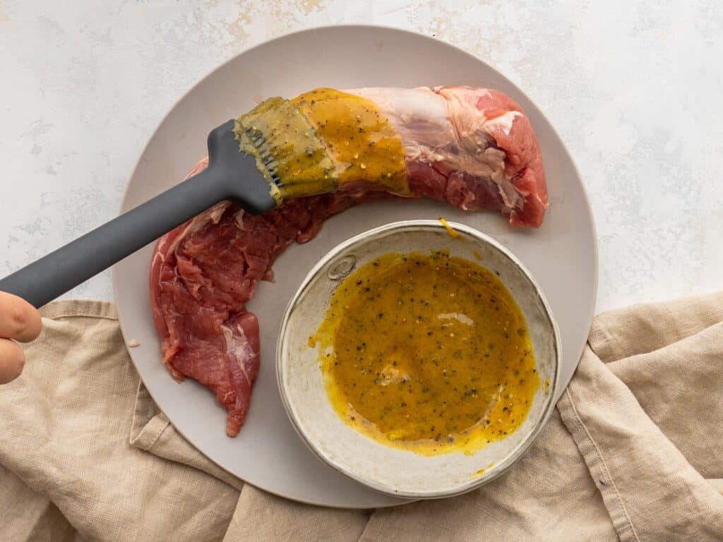 a basting brush brushing a brown sugar and dijon mustard marinade over a pork tenderloin