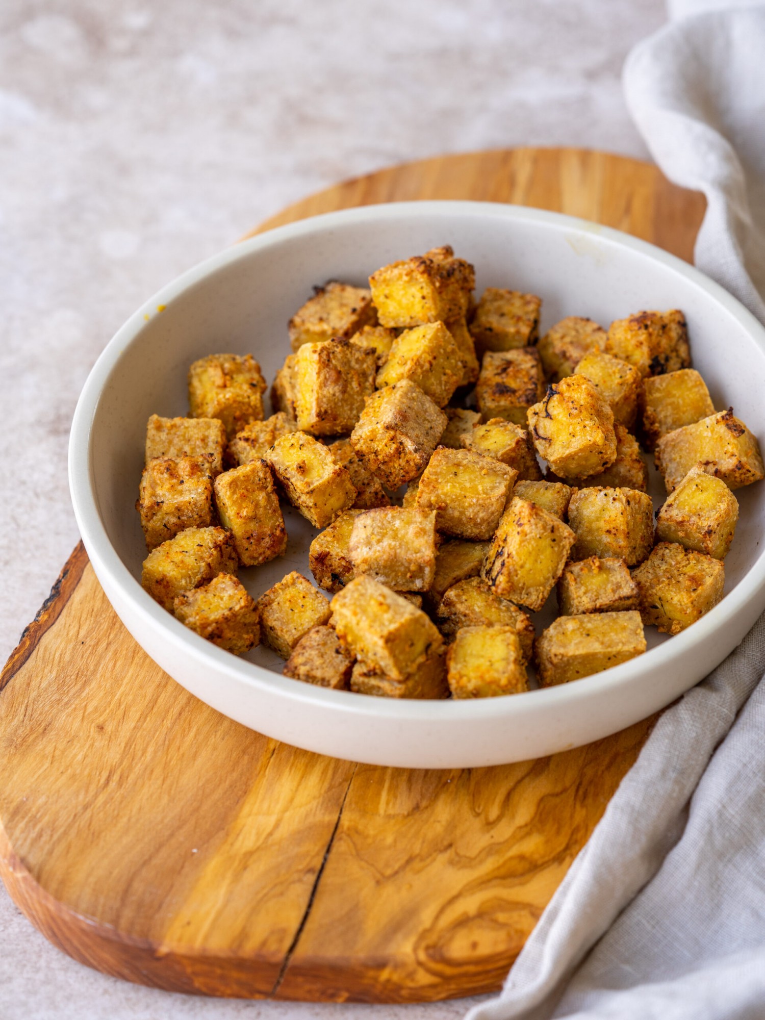 a three quarter view photo of a bowl of air fried tofu on a wooden cutting board