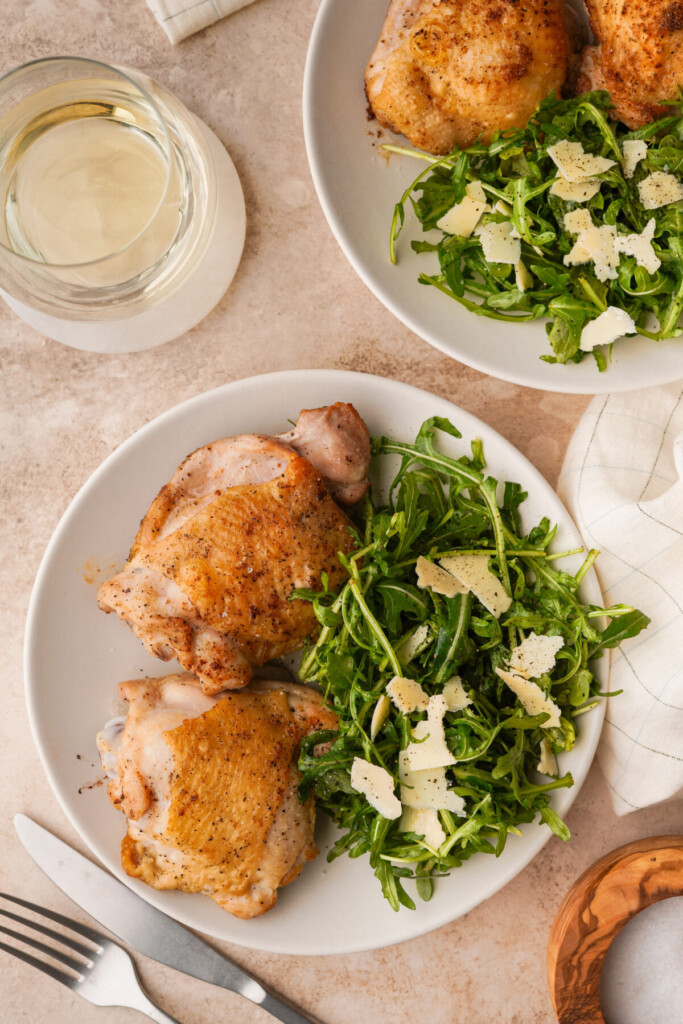 Seared chicken thighs n a serving plate with an arugula salad next to a glass of white wine