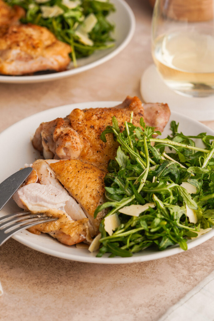 Three quarter view of a chicken thigh with a slice cut into one of the chicken thighs. There is an arugula salad on the plate with the chicken thighs