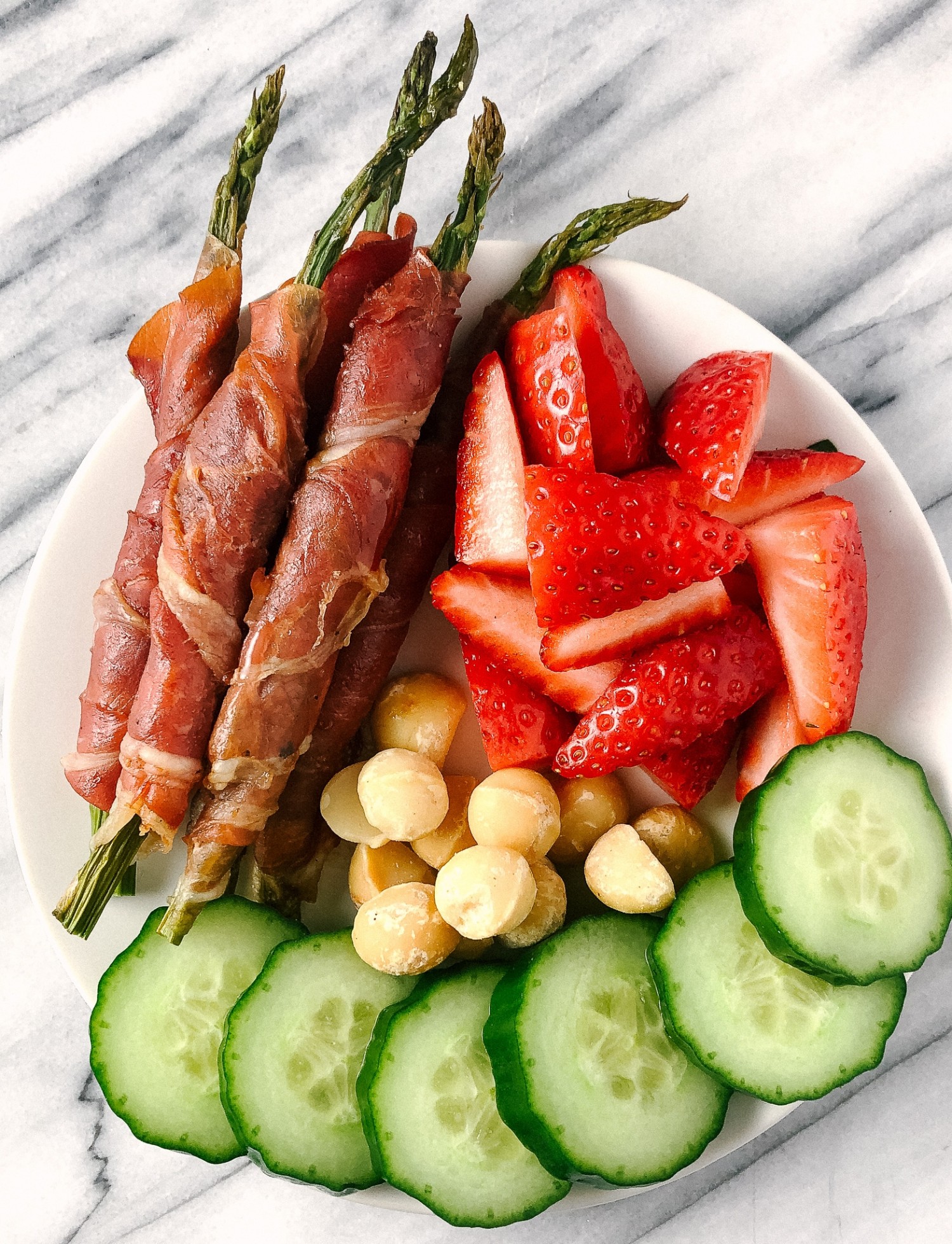 Above view of prosciutto wrapped asparagus on a snack plate with strawberries, macadamia nuts and sliced cucumbers