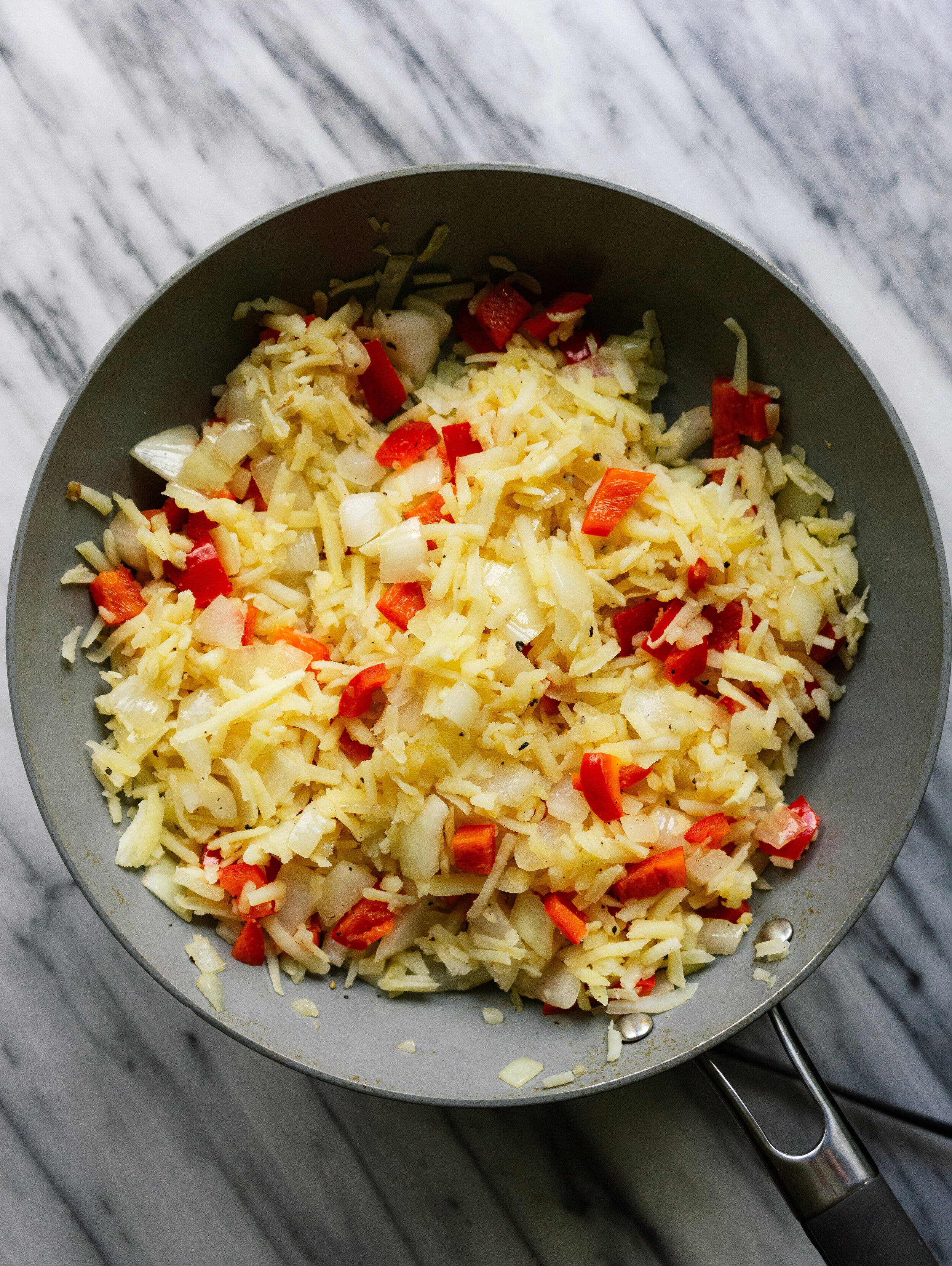 Above view of hash browns in a frying pan with onions and red bell peppers