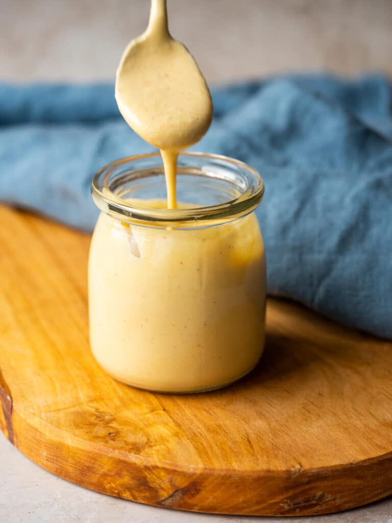 Side view of roasted garlic aioli dripping from a spoon into a jar of the sauce