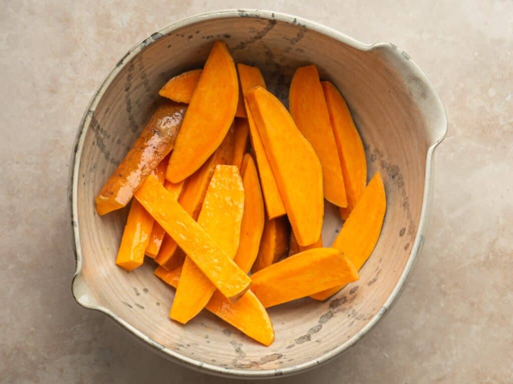 a large bowl filled with raw sweet potatoes cut into wedges and tossed in oil