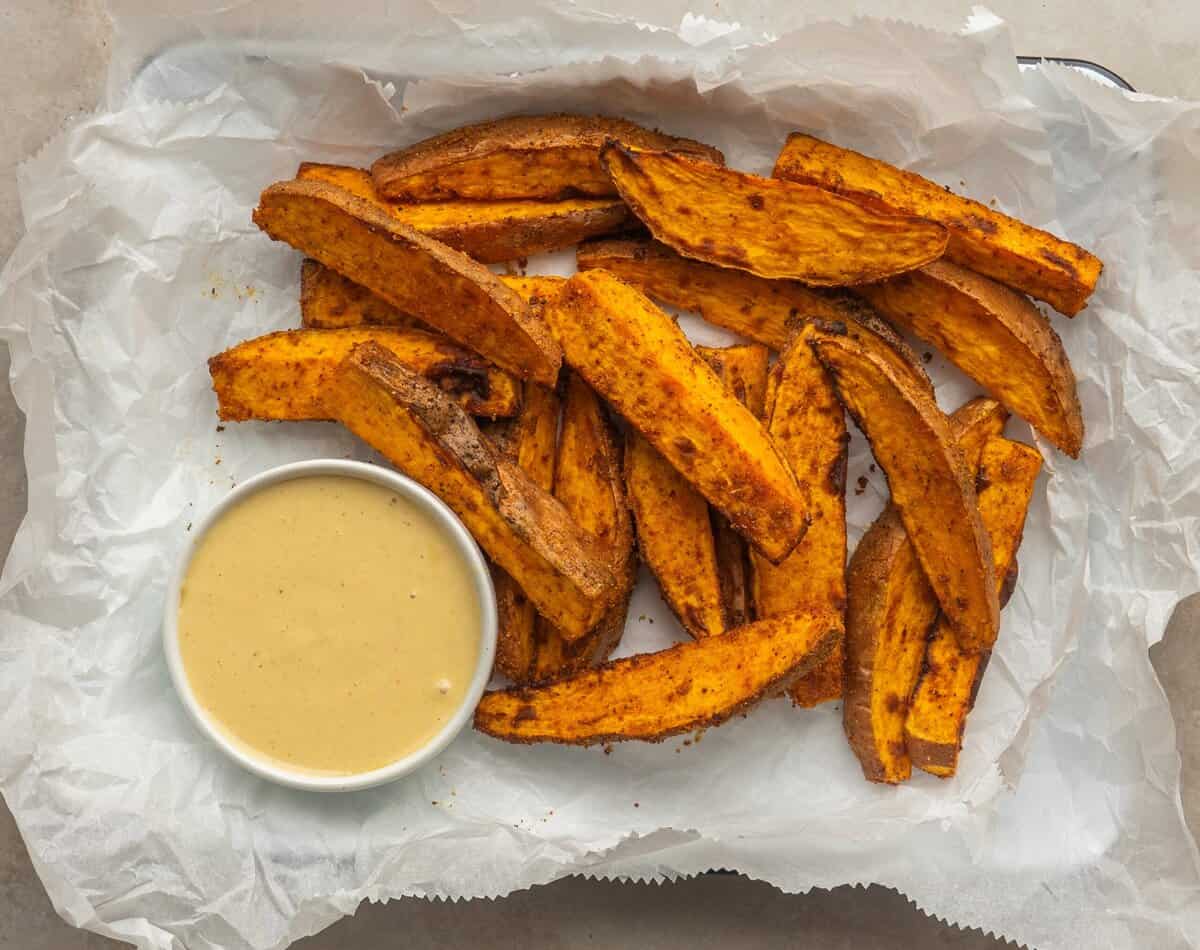 Air fried sweet potato wedges on a parchment lined tray with a little dish of dipping sauce next to it