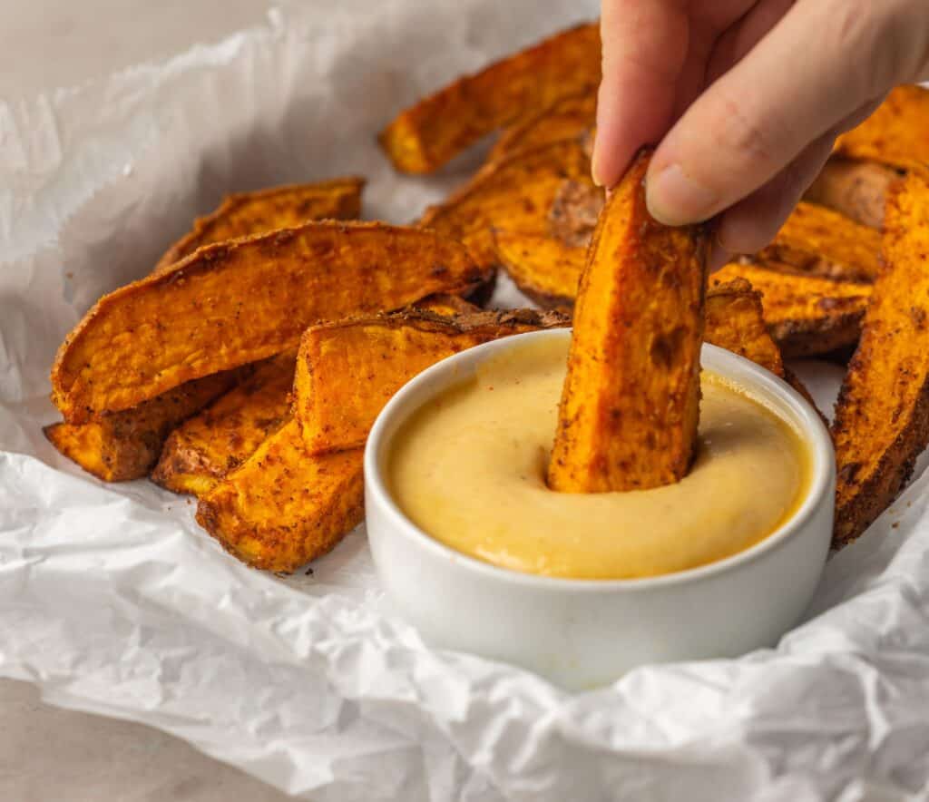 A photo of a hand holding an air fried sweet potato wedge into a cup of a dipping sauce