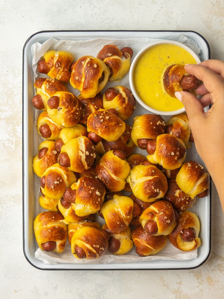 Above view of mini pretzel dogs on a serving platter being dipped into a homemade honey mustard