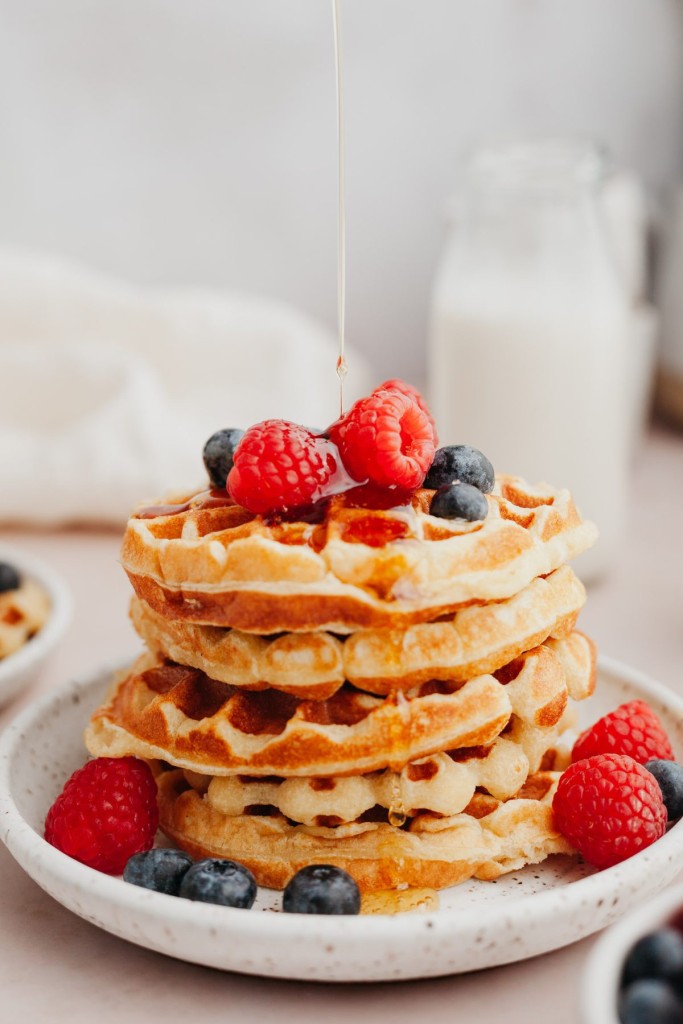 Side view of a stack of protein waffles with fresh berries on top and syrup pouring down onto the waffles
