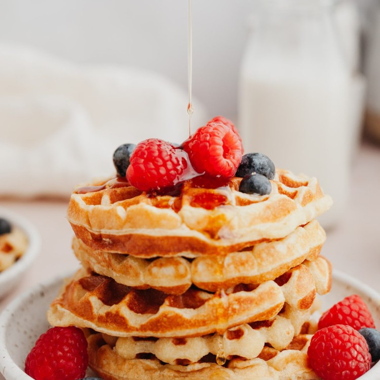 Side view of a stack of protein waffles with fresh berries on top and syrup pouring down onto the waffles
