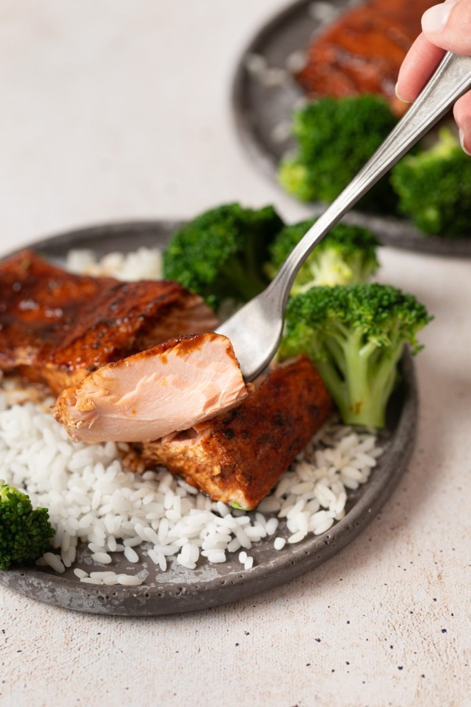 A fork breaking into a tender piece of honey glazed salmon on a plate with rice