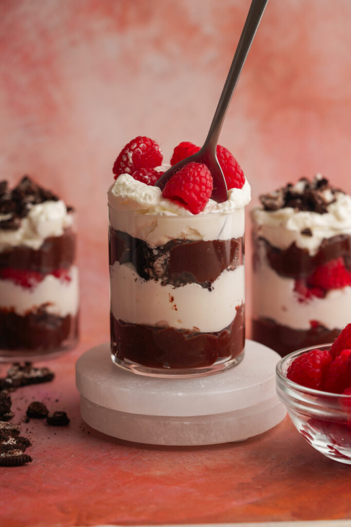 a side photo of chocolate parfait with raspberries on top with a spoon in it and two other parfaits in the backgroun