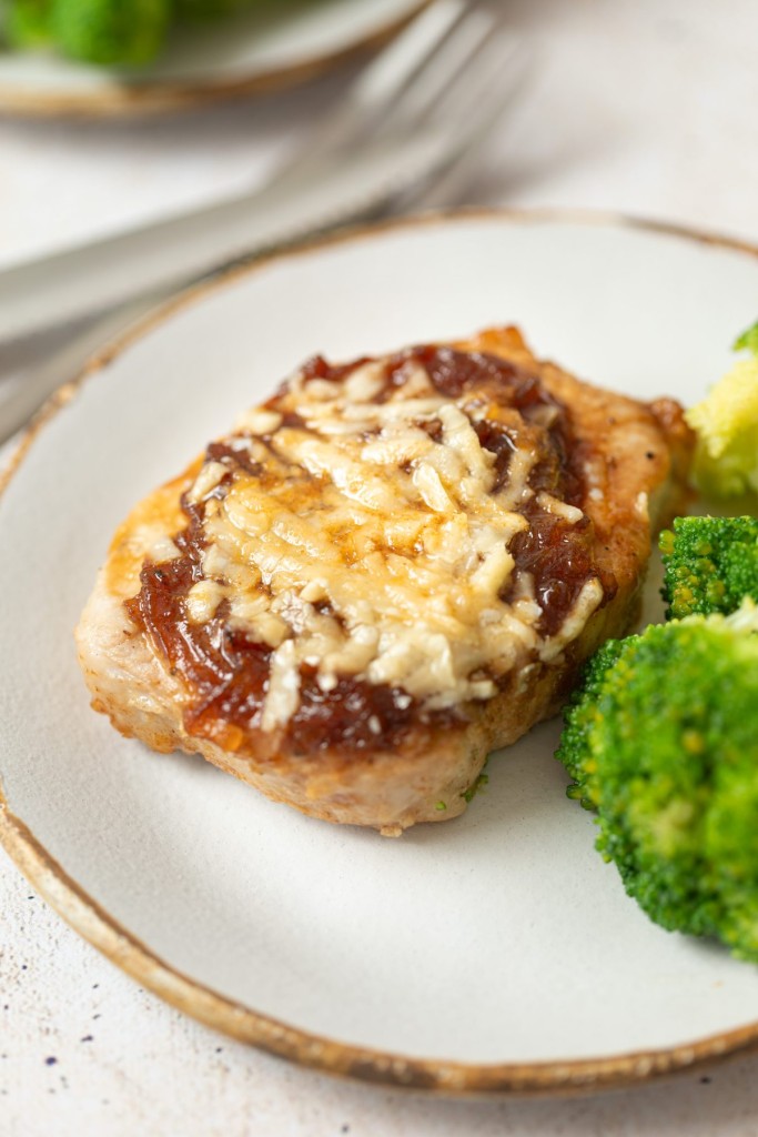 a three quarter photo of french onion pork chop on a white plate with broccoli next to it 