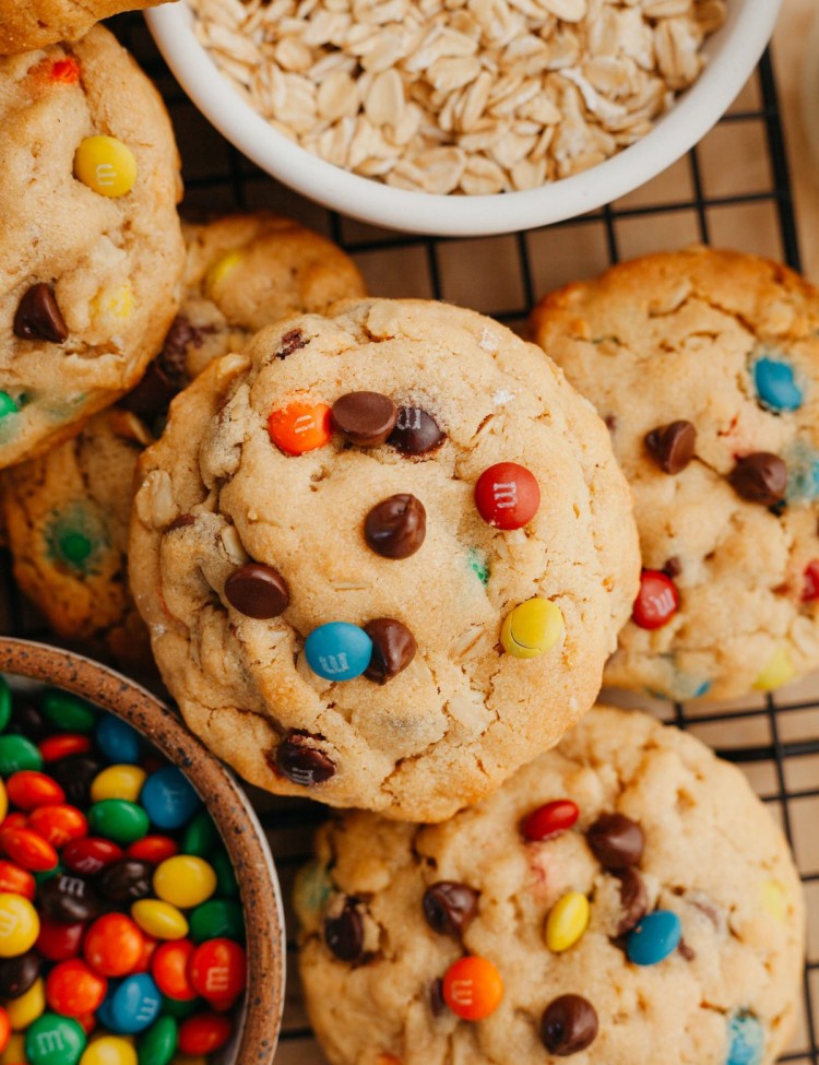 Close up above view of a bunch of monster cookies stacked up with one cookie on top. A small bowl of mini m&ms and oats are next to the cookies
