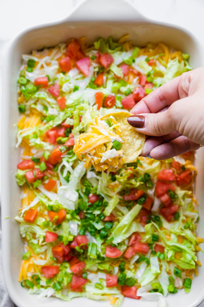 an above image of a hand holding a scoop of taco dip on a chip above a baking dish with taco dip