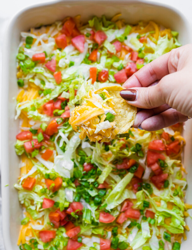 an above image of a hand holding a scoop of taco dip on a chip above a baking dish with taco dip